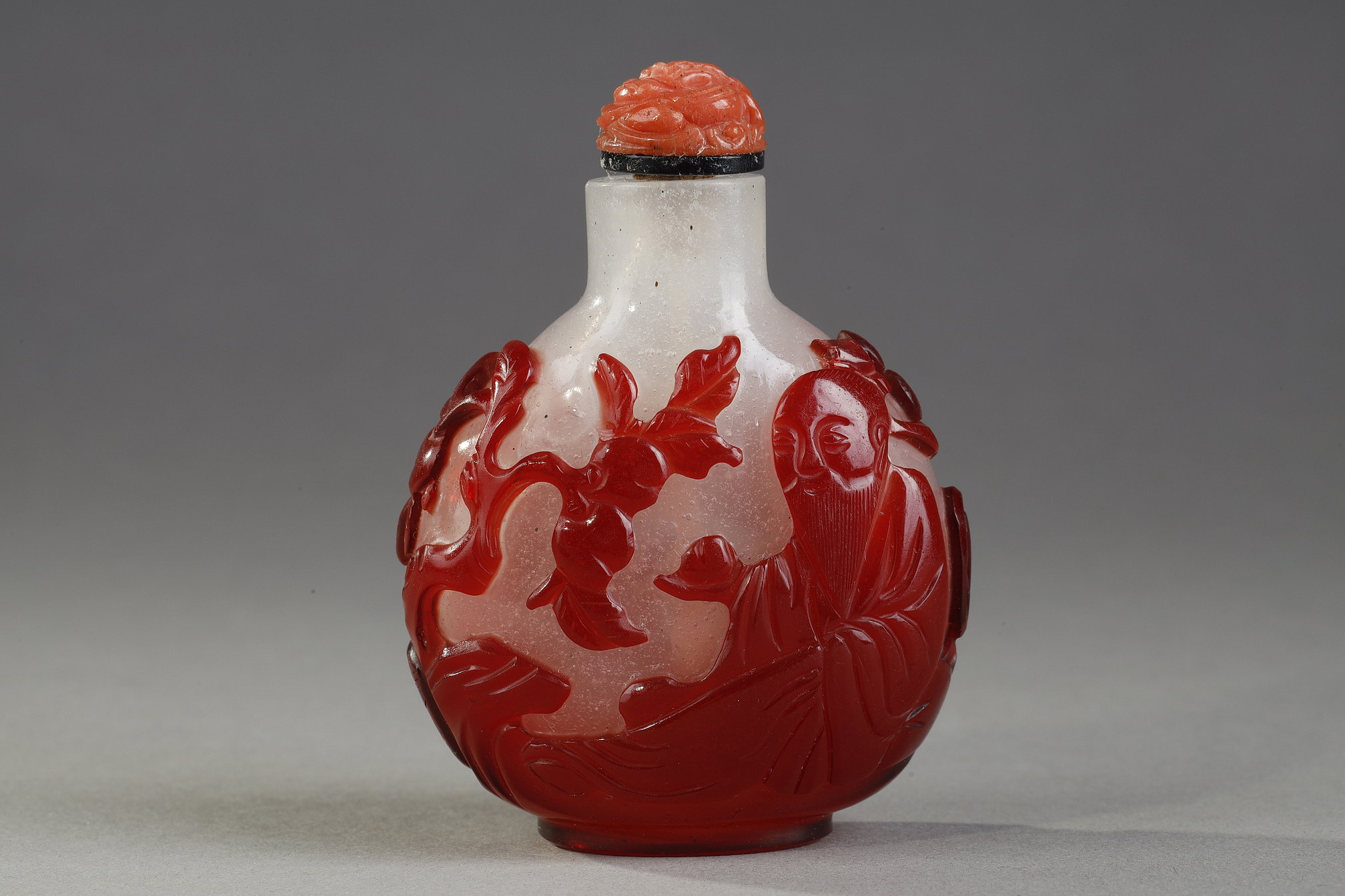 Snuff Bottles : Snuff bottle  red overlay glass with Shou Lao holding a peche of longevity and looking at peches and on the other side of a character holding a cup on a tray in front of a pine - China 1800/1850